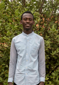sponsorship photo of a posed, smiling young man wearing a blue collared shirt, standing in front of green bushes